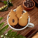 Mug with cookies shaped like pine trees and a small bowl of coffee beans on a wooden surface.