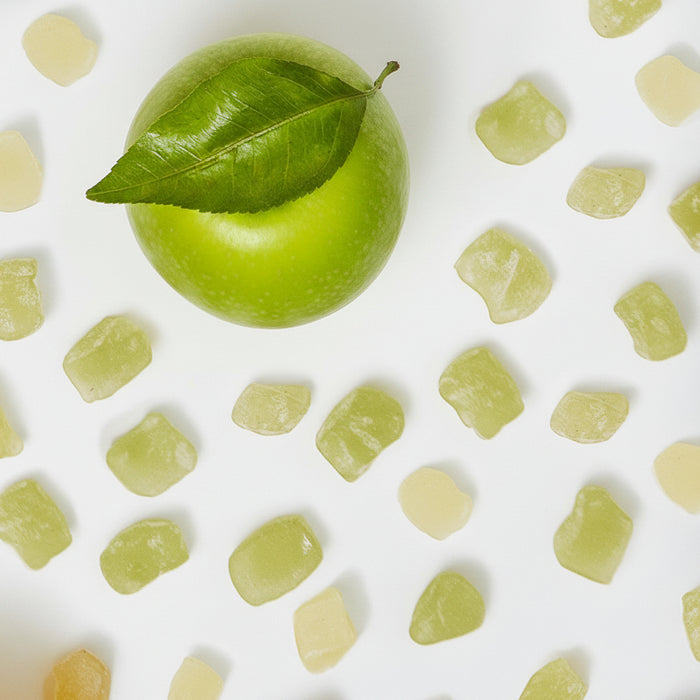 A green apple surrounded by small pieces of green mochi candy on a white background. 
