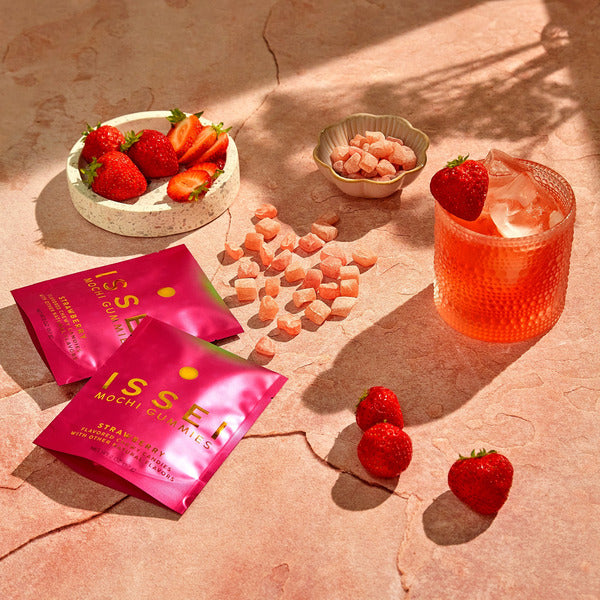 Two pink ISSSEL Mochi Gum Mils packets for strawberry flavor are displayed on a textured terracotta surface alongside fresh strawberries, loose mochi candies, a small bowl of dried strawberries, and a glass of strawberry iced drink.