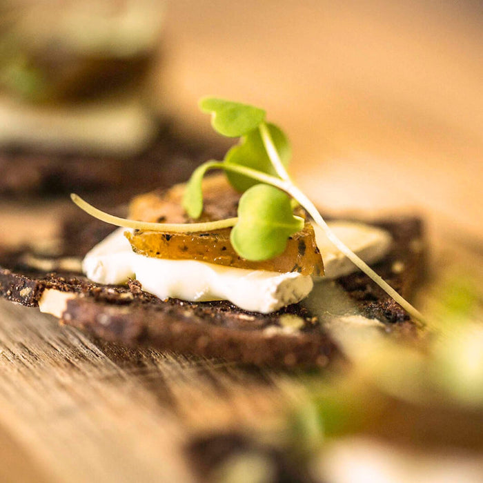 A close-up shot of a gourmet food item, featuring a dark cracker topped with a slice of white brie cheese, a slice of POG fruit paste, and fresh green microgreens, set on a wooden surface.