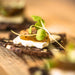A close-up shot of a gourmet food item, featuring a dark cracker topped with a slice of white brie cheese, a slice of POG fruit paste, and fresh green microgreens, set on a wooden surface.